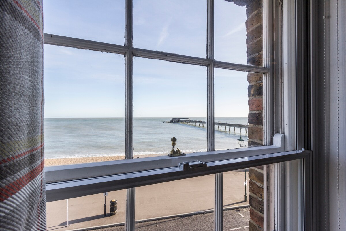 A view through a window showcases the English Channel, with a sandy beach and a pier extending into the water. The tranquil sea and clear sky create a serene backdrop, framed by the window's classic design featuring wooden panes.