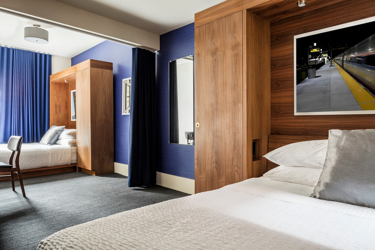 A spacious guestroom features a large bed with soft pillows and a gray throw. Opposite, a second sleeping area contains a single bed. Warm wooden cabinetry complements the blue accent walls, while large curtains frame a window, allowing natural light to brighten the space.