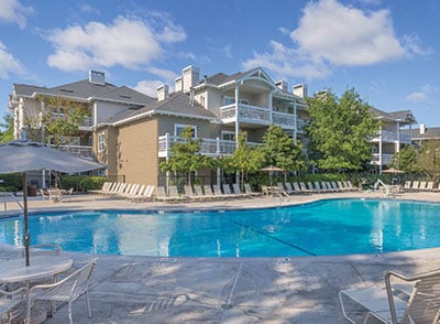 A spacious outdoor pool is surrounded by loungers and shaded areas. The three-story resort buildings are visible in the background, featuring balconies and lush greenery. Clear blue skies and soft clouds enhance the inviting atmosphere of the pool area.