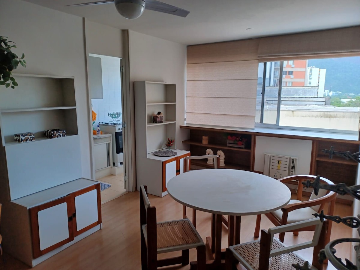 A bright living area features a round dining table surrounded by four wooden chairs. Shelving units are positioned against two walls, extending natural light through large windows with light-filtering shades. A view of the outdoors is visible beyond the window, enhancing the open space.