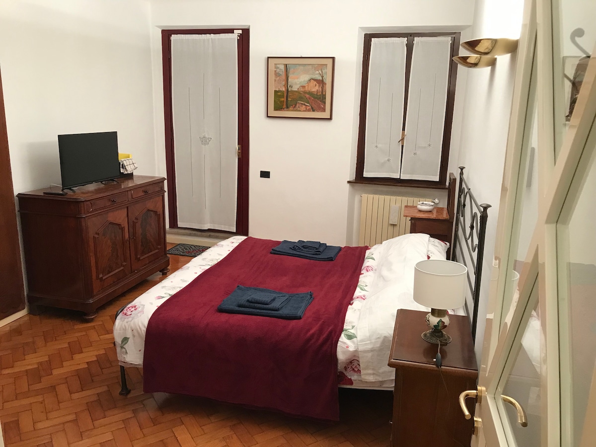 A comfortable room features a spacious bed adorned with a deep red blanket and two navy towels. A wooden wardrobe and a bedside table are visible, along with a television. Two windows provide natural light, and artwork is displayed on the wall.