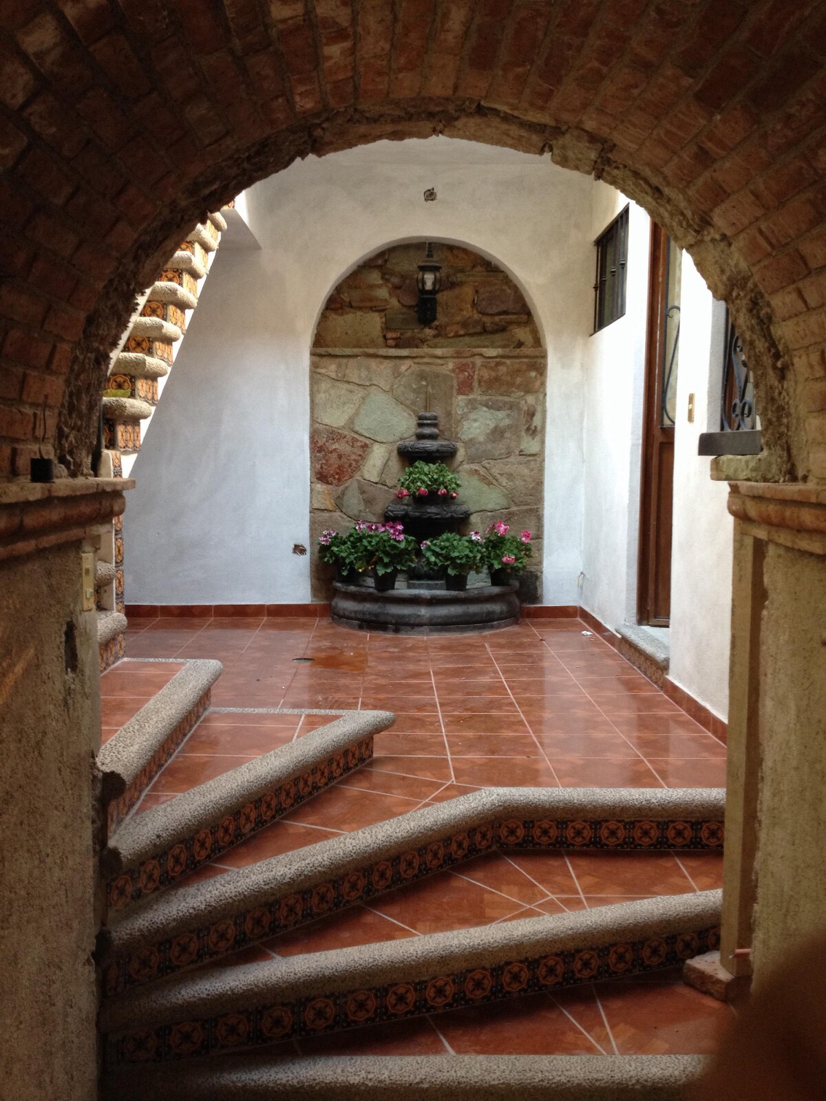 An arched entrance frames a serene courtyard featuring a small stone fountain surrounded by vibrant flowers. A staircase with carefully designed steps leads up, while the terracotta tiled floor reflects natural light flowing through nearby windows.