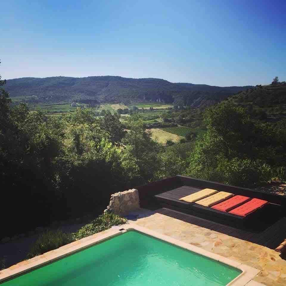 A private swimming pool is positioned in the foreground, offering refreshing green water. Surrounding greenery enhances the scenic view of rolling hills and valleys in the distance. Sun loungers in various colors are arranged nearby, providing space to relax and enjoy the natural surroundings.