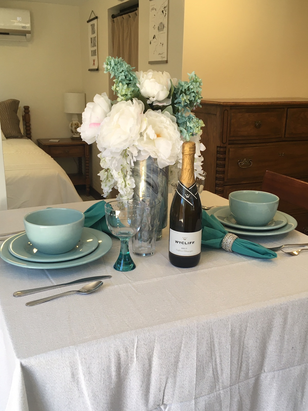 A dining table is set for two, featuring soft blue dishware and sparkling glassware. A bouquet of white flowers is arranged in a clear vase, enhancing the serene ambiance. A bottle of champagne is placed at the center, with a tasteful linen tablecloth draped beneath.