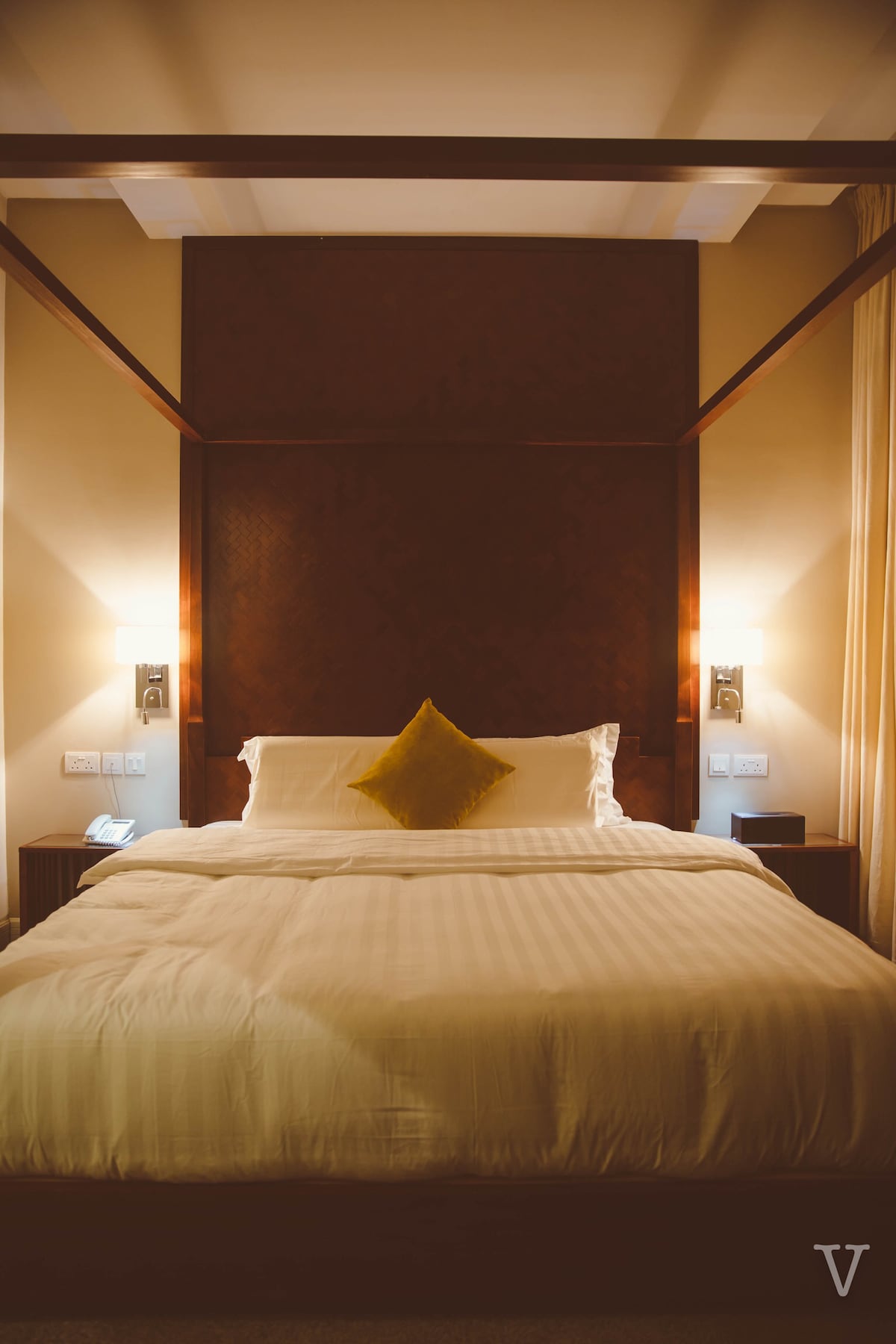 A spacious bed is centrally positioned against a dark wood headboard, with soft white bedding neatly arranged. Two bedside lamps provide warm illumination, and a decorative yellow cushion adds a pop of color. Light filters through sheer curtains, casting a gentle glow.