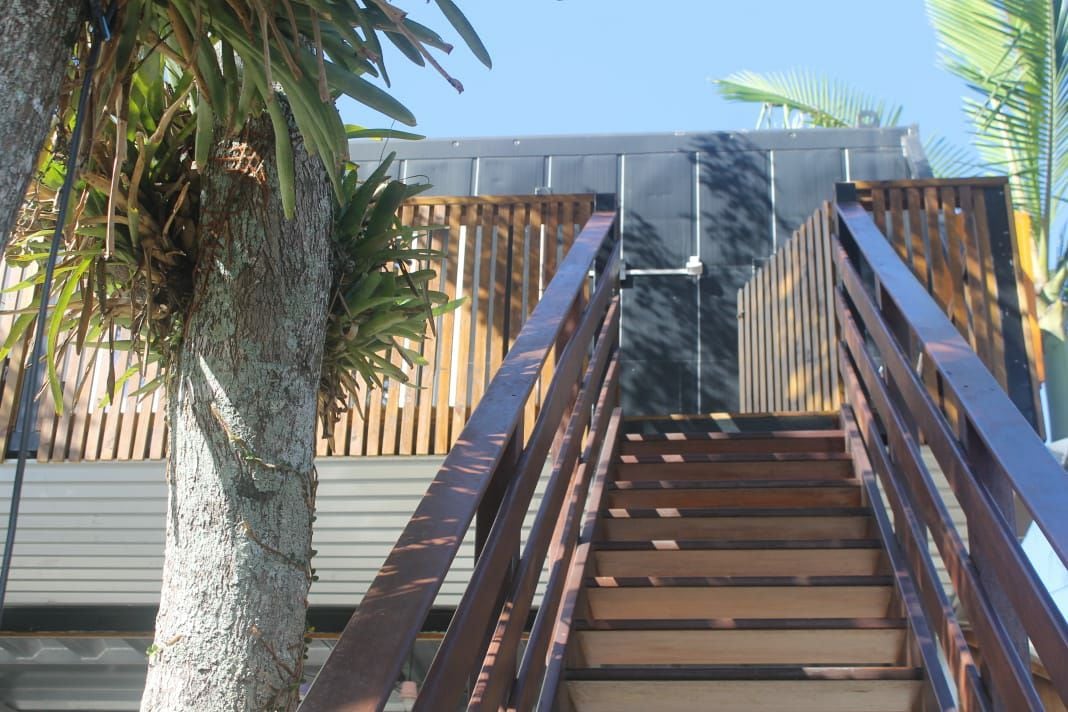 A set of wooden stairs ascends towards the entrance, framed by tall palm trees. The sturdy railing complements the modern design and leading structure, creating a welcoming approach to the upper level of the property.