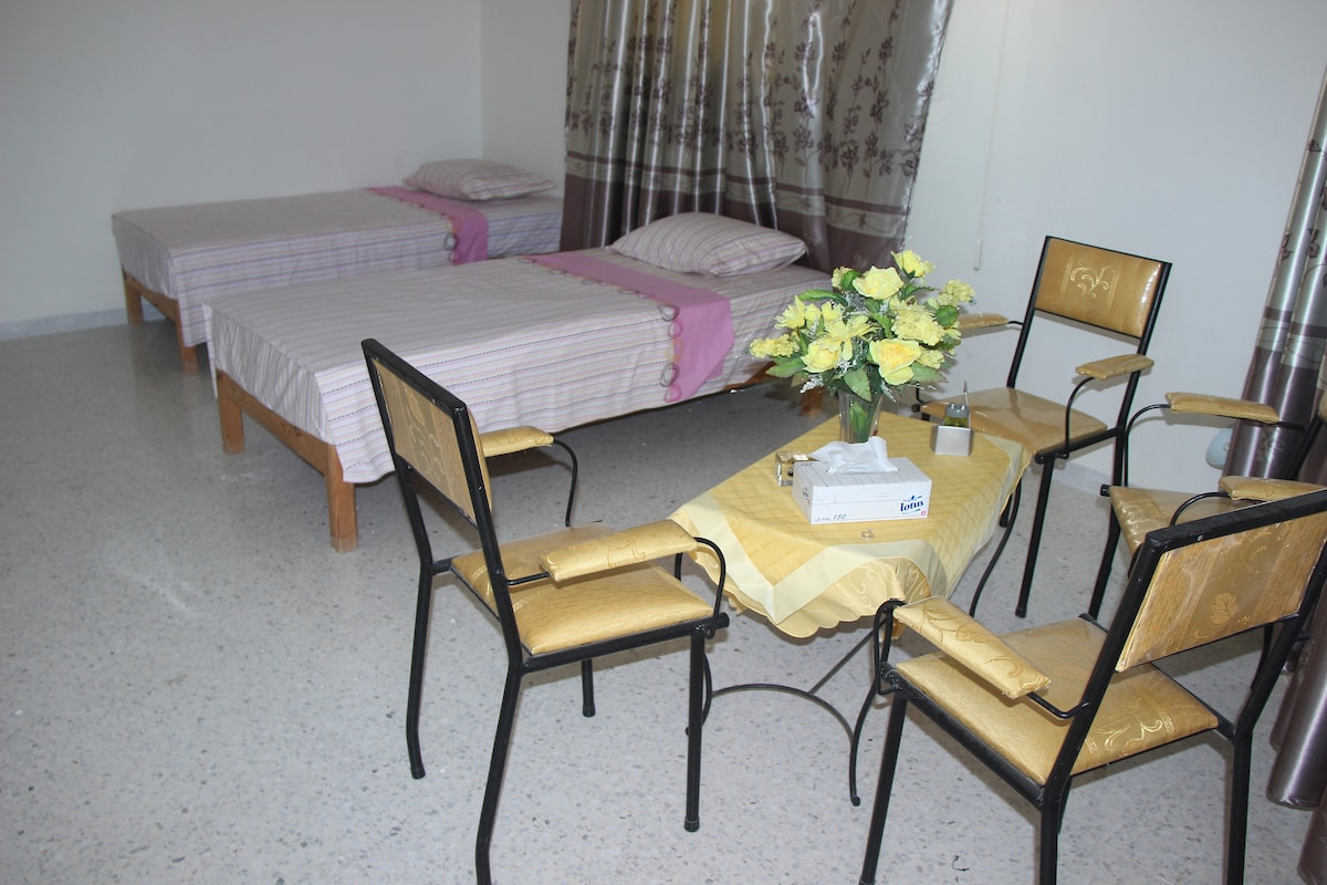 The room features two separate beds positioned next to each other, each made with pastel-colored bedding. A small round table is accompanied by four chairs, creating a cozy seating area. Light-colored curtains drape over the window, allowing natural light to fill the space.