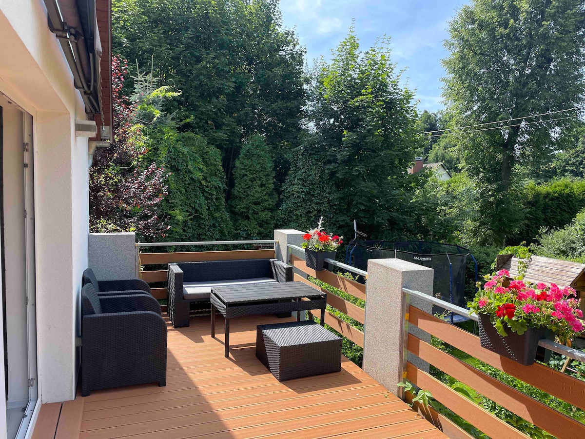 A private terrace is furnished with a seating area consisting of a dark wicker sofa set and a coffee table. Flower boxes filled with colorful blooms add greenery, while the surrounding foliage provides a natural backdrop.