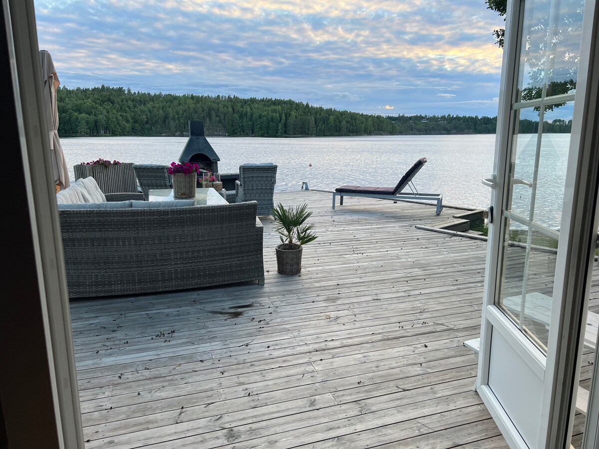 A spacious wooden deck overlooks a serene body of water, framed by lush greenery. Comfortable outdoor seating is present, along with a lounge chair and a potted plant. A softly colored sky is reflected in the water, enhancing the tranquil setting.