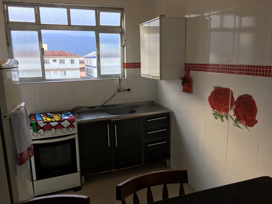 Cozinha com boa ventilação, geladeira, gabinete e pia, fogão com botijão de gás, armários suspensos, pratos, copos e talheres.