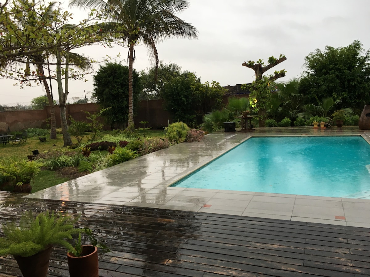 A modern pool area features a clear blue pool surrounded by tropical plants and trees. The deck is made of wooden planks, and rain is visible on the surface, creating a serene atmosphere. Lush greenery lines the perimeter, enhancing the tranquil setting.
