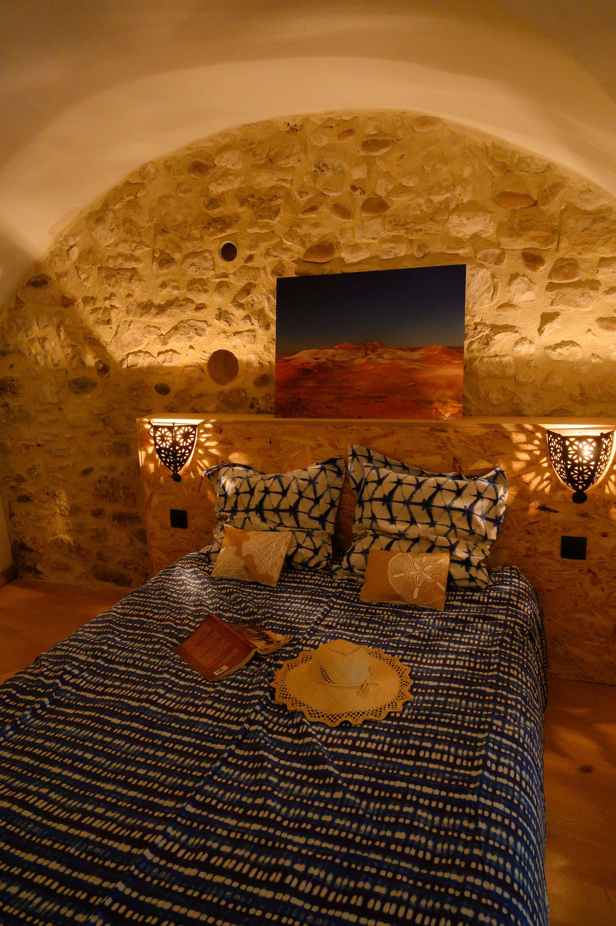 The sleeping area features a spacious bed with a patterned blue and white coverlet, accented by decorative pillows. The stone wall in the background adds texture, while two lanterns provide soft illumination. A piece of artwork depicting a desert landscape is displayed above the headboard.