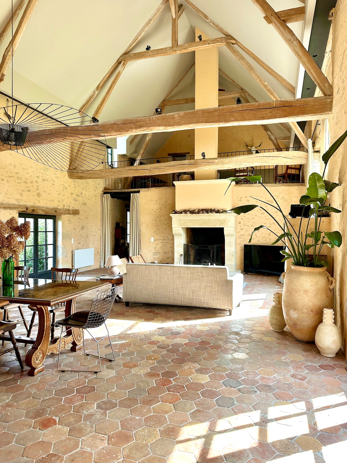 A spacious living area is highlighted by high ceilings with exposed beams. A neutral-toned sofa faces a fireplace, while a dining table with chairs occupies one side. Natural light enters through large windows, illuminating the warm terracotta floor and decorative plants.