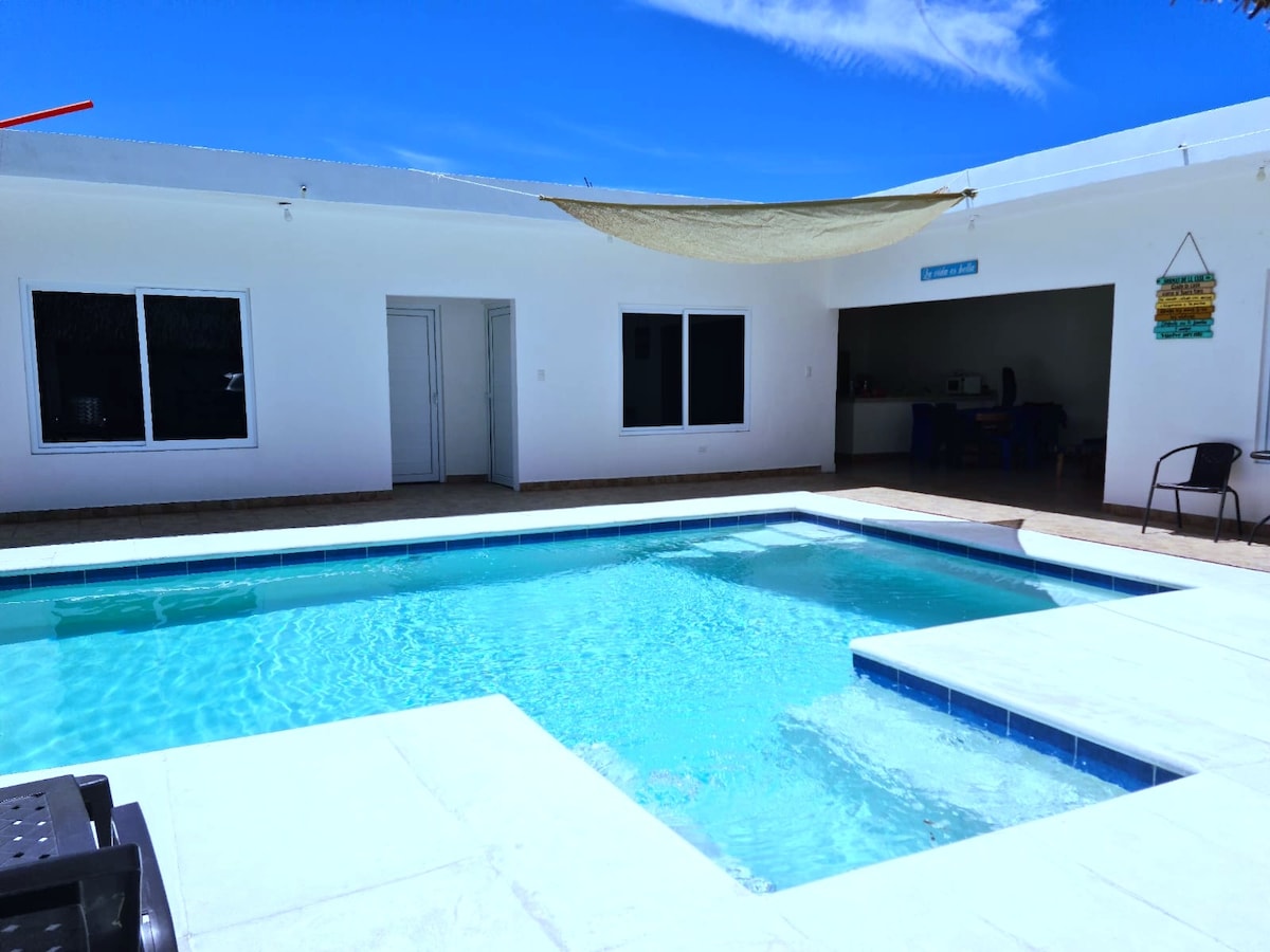 A refreshing swimming pool is centrally located, surrounded by a spacious patio. A shaded area is provided by a canopy above, while large windows reveal the interior space, enhancing natural light. The pool is designed for relaxation and enjoyment.
