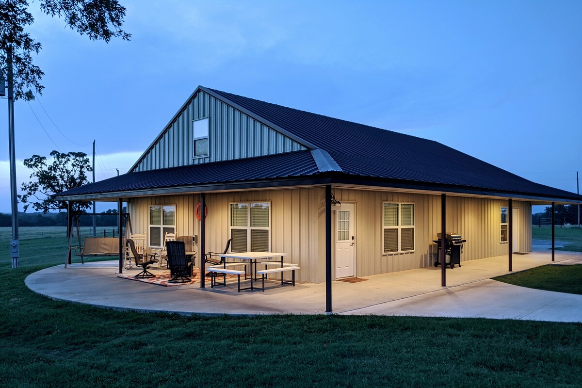 A spacious building is set in a serene rural landscape, with a covered porch featuring outdoor seating. Large windows allow natural light to illuminate the exterior. The surrounding green lawn and open fields contribute to the tranquil atmosphere, especially during the evening hours.