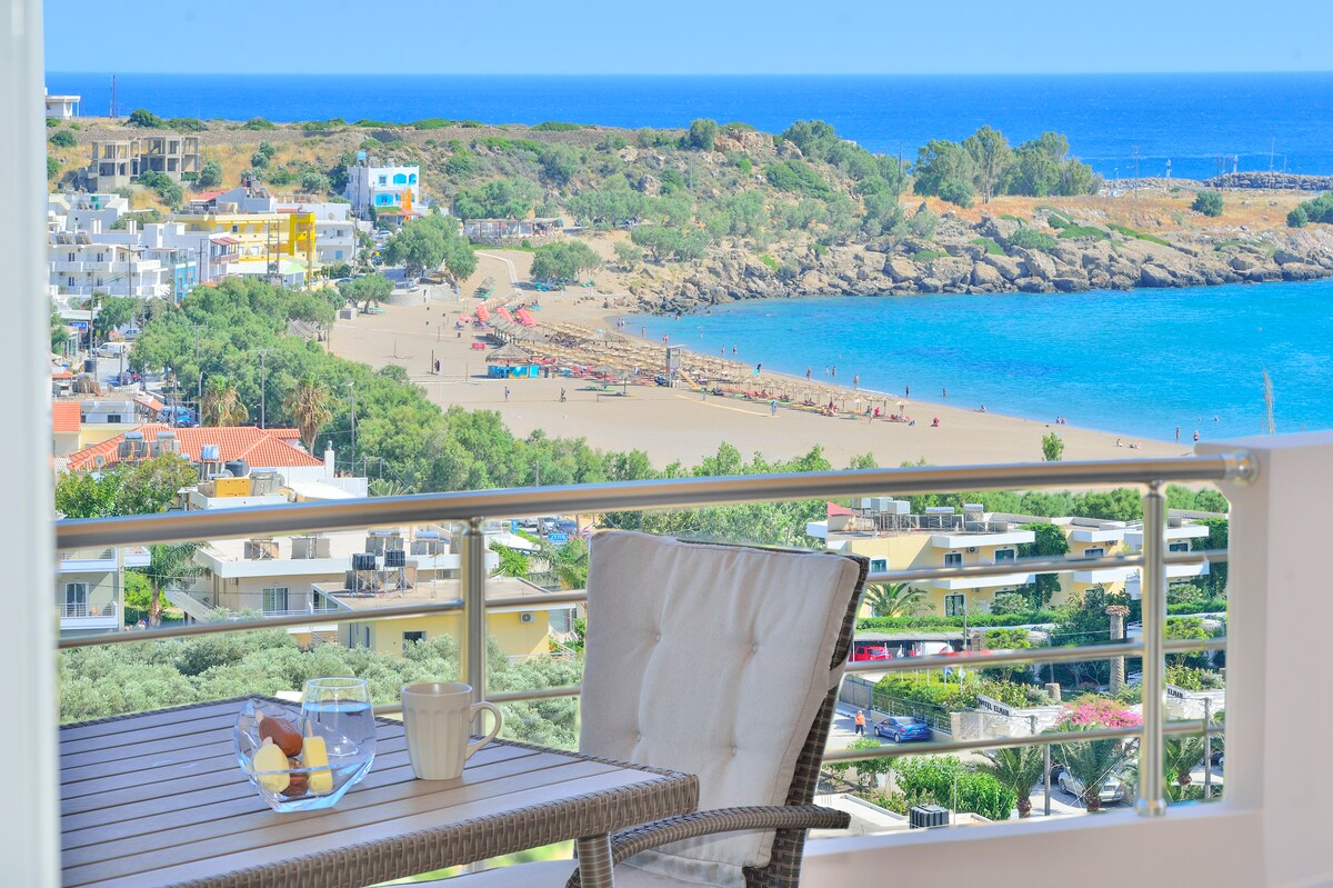 A balcony table set for two overlooks a sandy beach and clear blue waters of the Mediterranean Sea. Colorful beach umbrellas dot the shore, while a coastal village is visible in the background, surrounded by lush greenery and rocky formations.