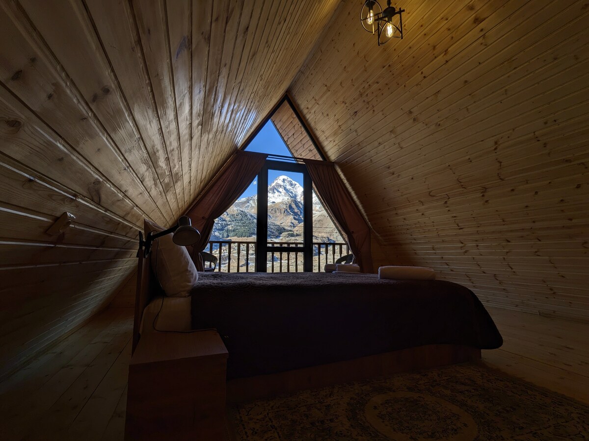 A cozy sleeping area features a spacious bed with neutral bedding, positioned against angled wooden walls. Large windows frame a scenic mountain view, while curtains gently filter incoming light. A bedside lamp and a rolled towel are visible, adding to the inviting ambiance.