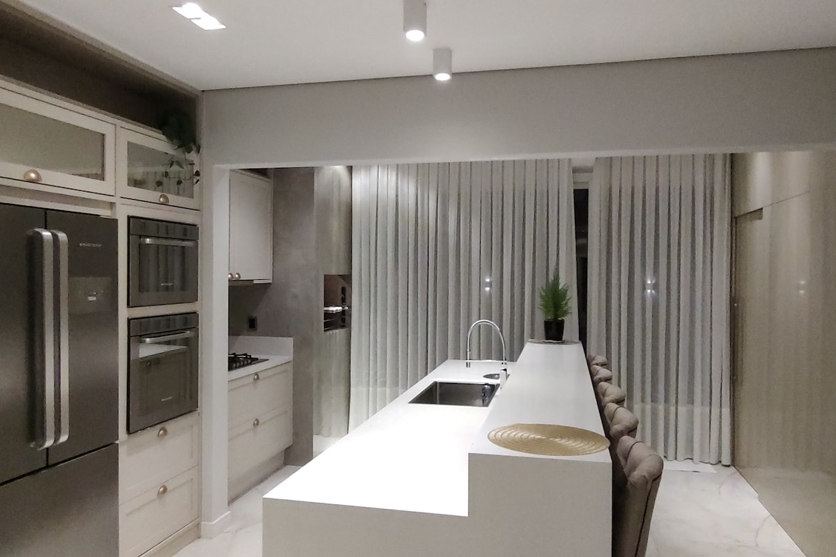 A modern kitchen features sleek cabinetry with built-in ovens and an expansive white countertop. Ample seating is arranged along the counter, while soft drapes cover large windows, allowing gentle light to fill the space. A potted plant adds a touch of greenery.