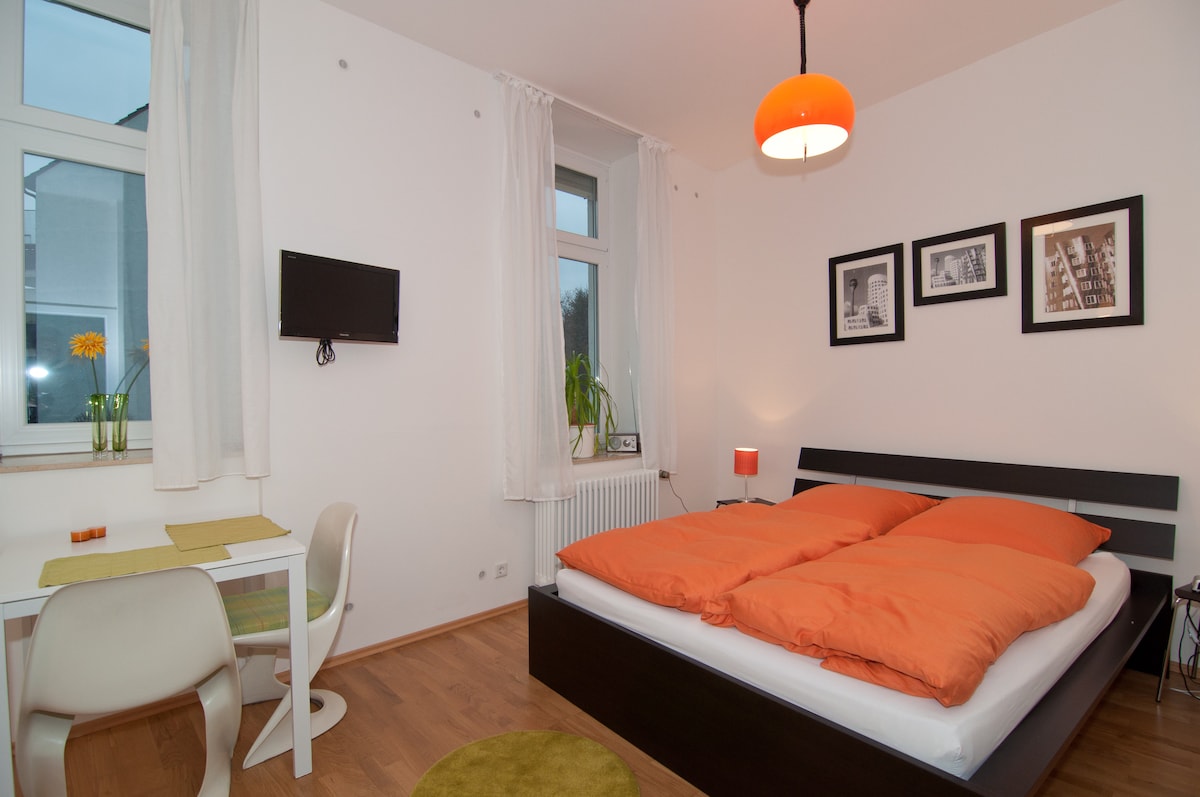 A bright sleeping area is shown, featuring a spacious bed with orange bedding and a minimalist design. A small dining table with two chairs is present, alongside a flat-screen TV mounted on the wall. Large windows allow natural light to enter the room.