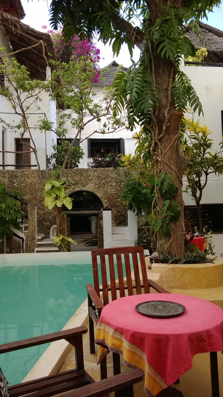 A serene courtyard features a clear turquoise pool surrounded by lush greenery and vibrant flowers. A wooden table is set with a patterned cloth, accompanied by a pair of rustic chairs. The tranquil scene is enhanced by the natural texture of the stone wall in the background.