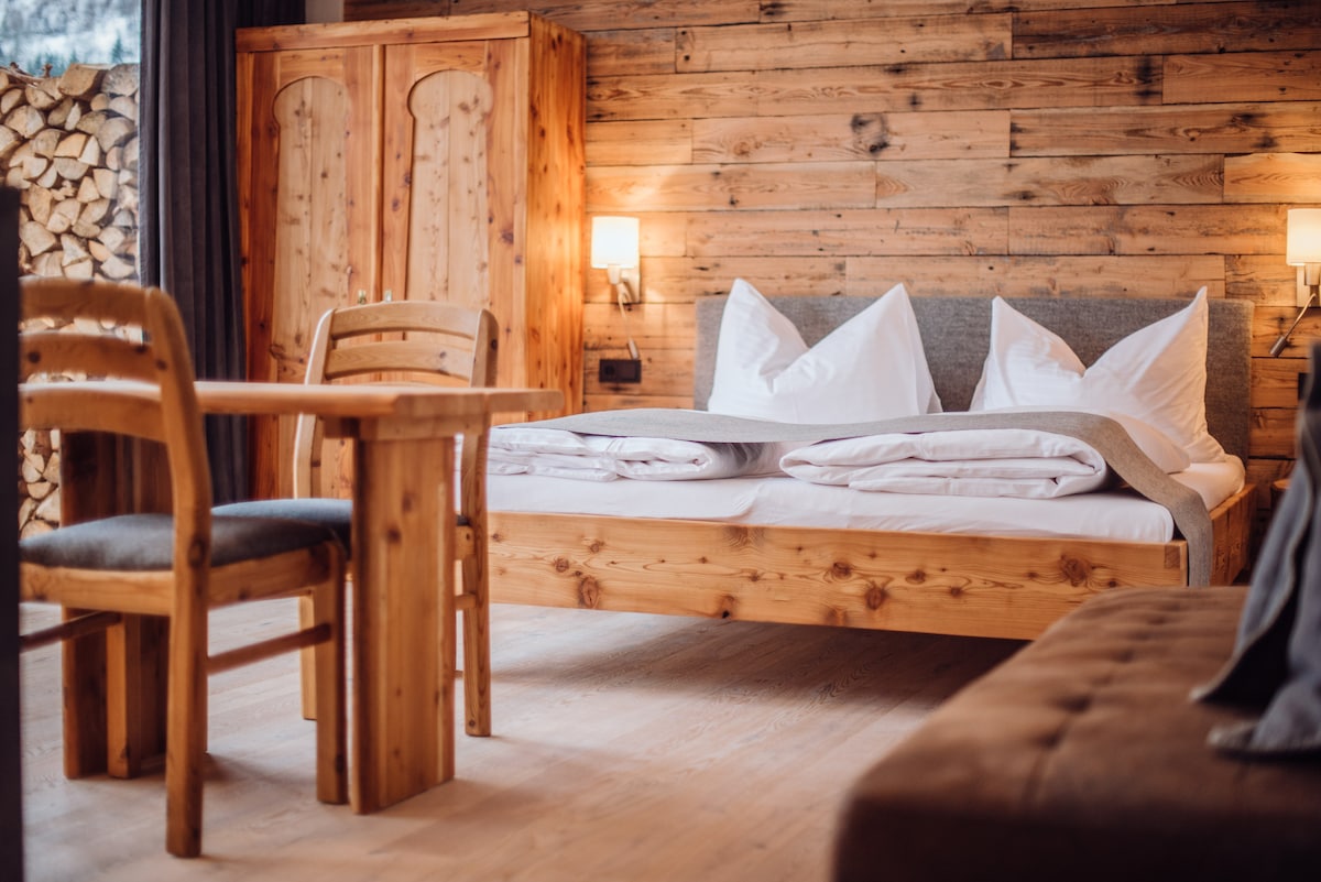 The room features a spacious wooden bed adorned with neatly arranged white linens and multiple pillows. A cozy wooden table and chairs complement the rustic wooden walls, while soft lighting from sconces creates a warm ambience.
