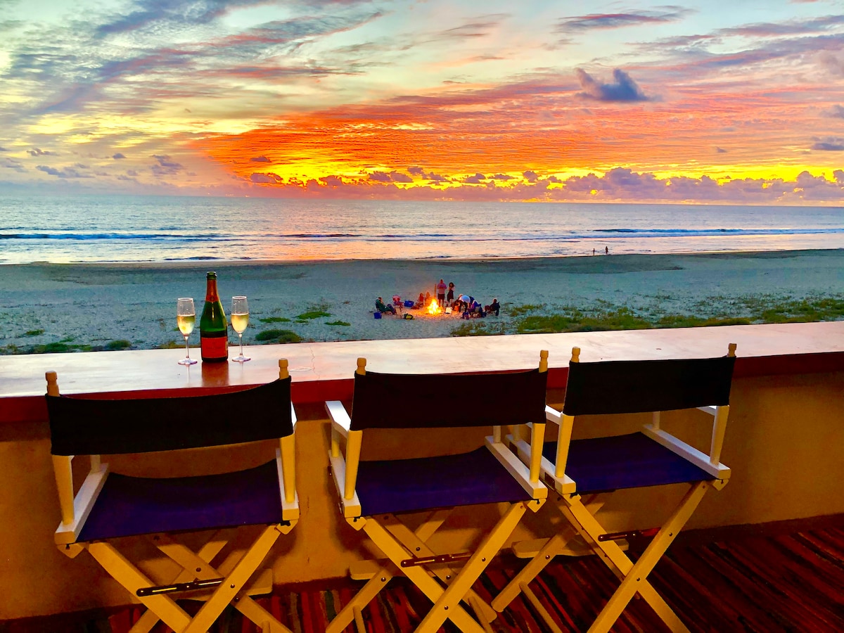 Three chairs are positioned on the balcony, offering an unobstructed view of a colorful sunset over the ocean. A group can be seen gathered around a fire pit on the beach, with soft waves lapping at the shore in the background.
