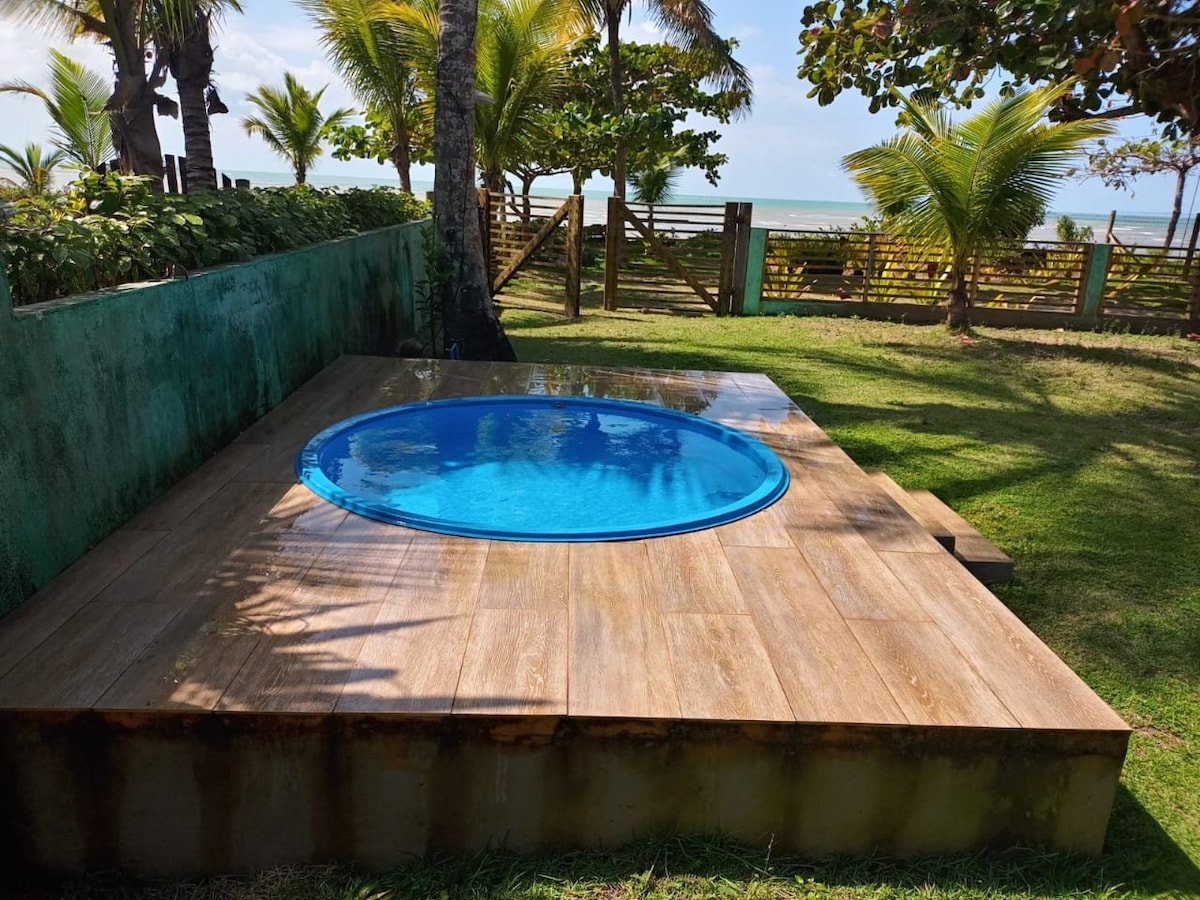 A circular pool is set on a wooden platform within a green lawn area. Surrounding palm trees provide a tropical ambiance, while the ocean is visible in the background beyond a wooden fence. The sun reflects off the water's surface.