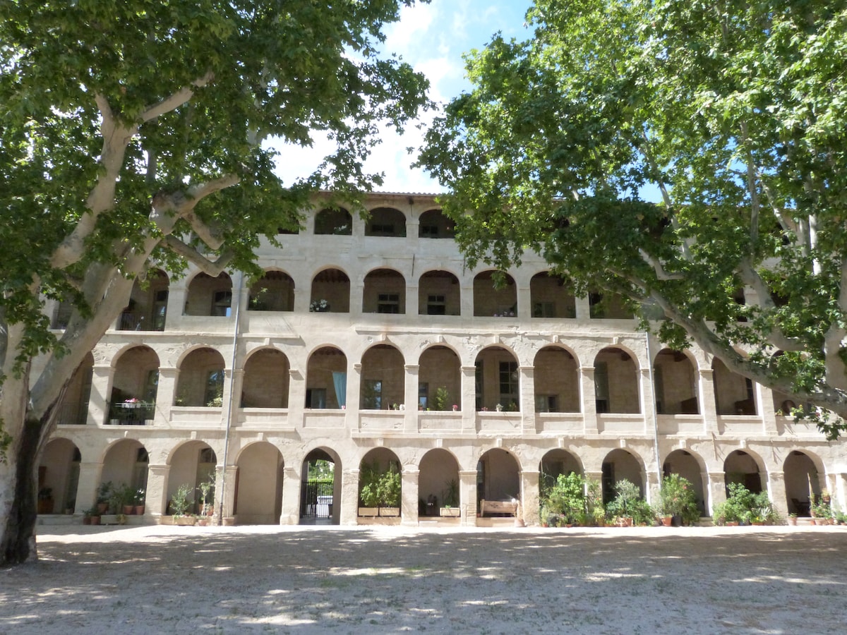 The historic building features an arched facade with multiple levels, framed by tall trees. An open courtyard is visible, showcasing greenery and potted plants. Natural light illuminates the stone structure, highlighting its architectural details.