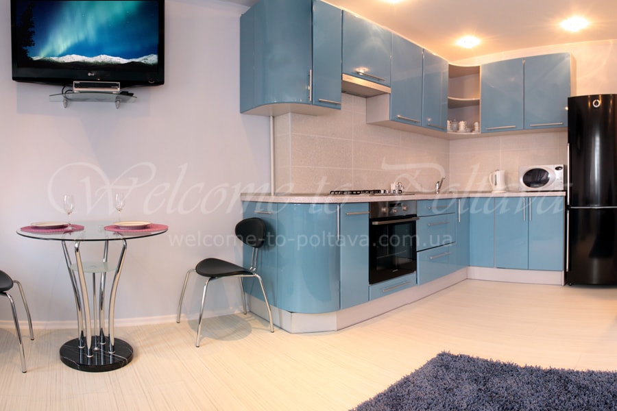 The kitchen area features sleek blue cabinetry with stainless steel appliances, including an oven and microwave. A round glass dining table is set with two chairs, situated near a black refrigerator. Soft lighting illuminates the space, enhancing the modern design.