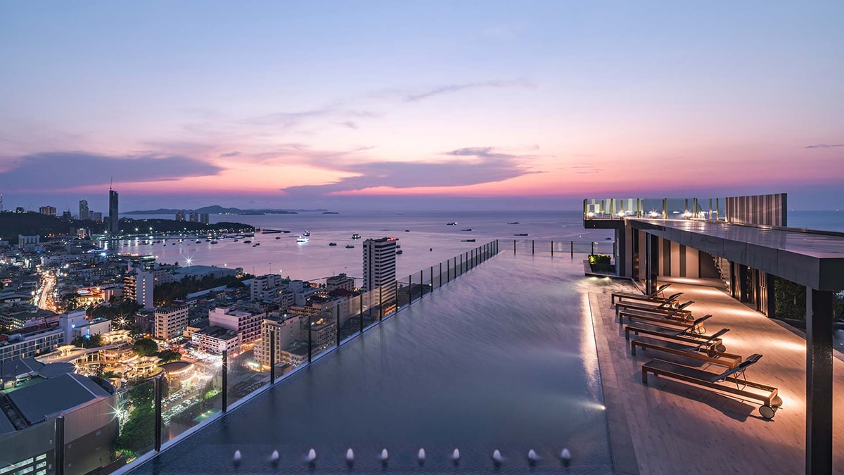 An expansive rooftop swimming pool offers stunning views of the ocean and city at sunset. Lounge chairs are positioned along the edge, inviting relaxation. The sky displays soft hues of pink and orange, reflecting the tranquil atmosphere of the setting.
