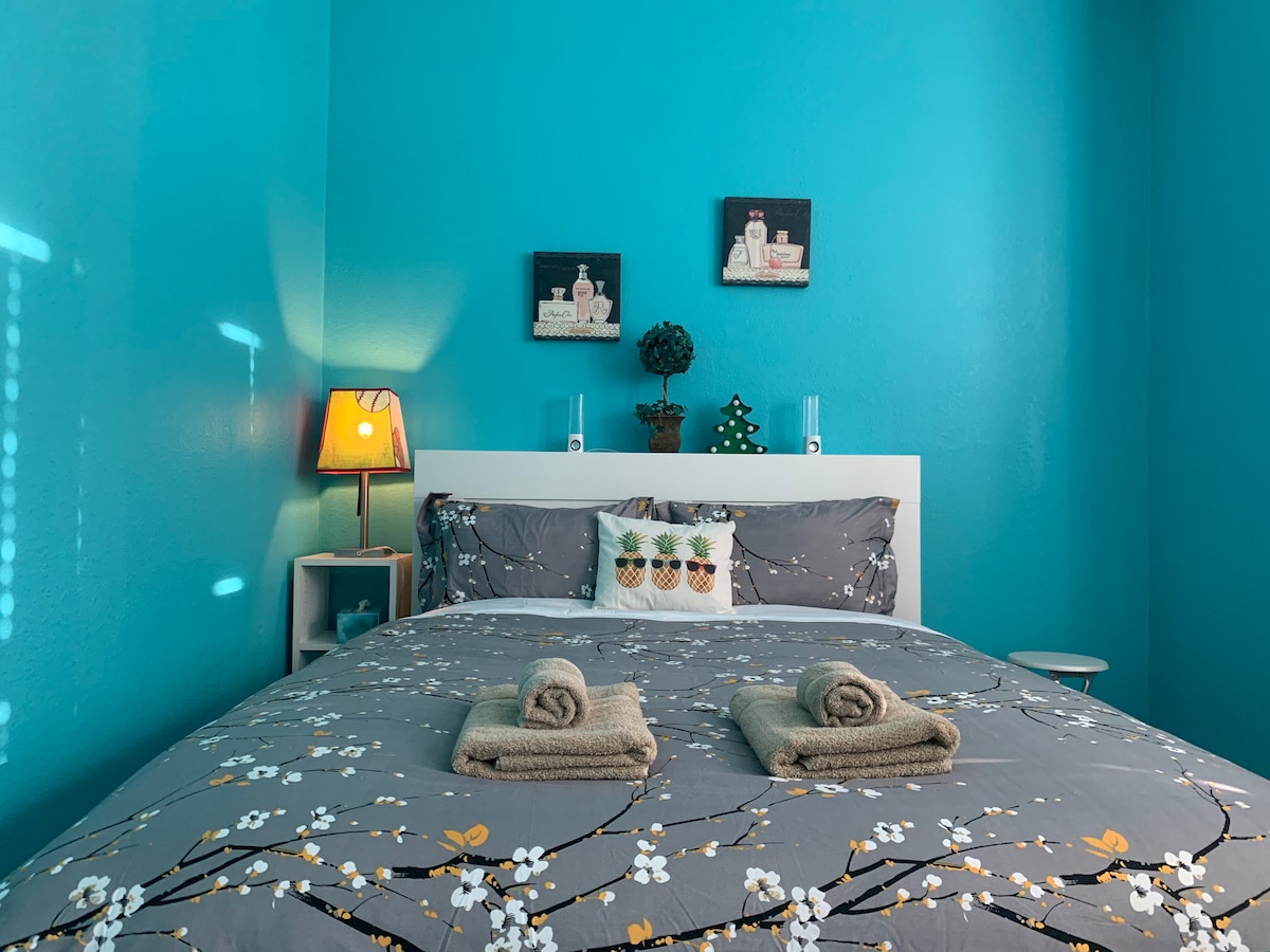 A vibrant sleeping area features a spacious bed with a floral-patterned duvet cover. Two rolled towels are neatly placed on the bed. Light turquoise walls are complemented by wall art and a small bedside lamp, providing a cozy atmosphere.