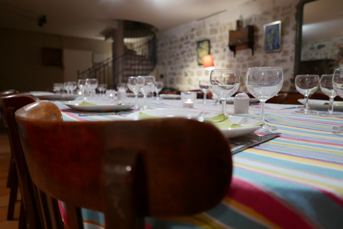 A long dining table is set with plates and glasses, adorned with a striped tablecloth. The room is illuminated softly, highlighting exposed stone walls and a spiral staircase in the background, creating a welcoming setting for communal dining.