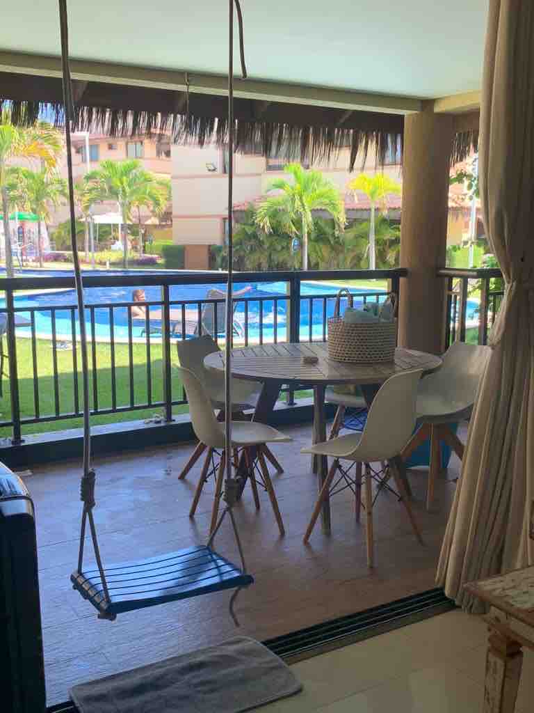 A covered balcony features a round dining table surrounded by four chairs, with a swing hanging nearby. Lush greenery and a tranquil pool are visible just beyond the railing, creating a peaceful outdoor setting.