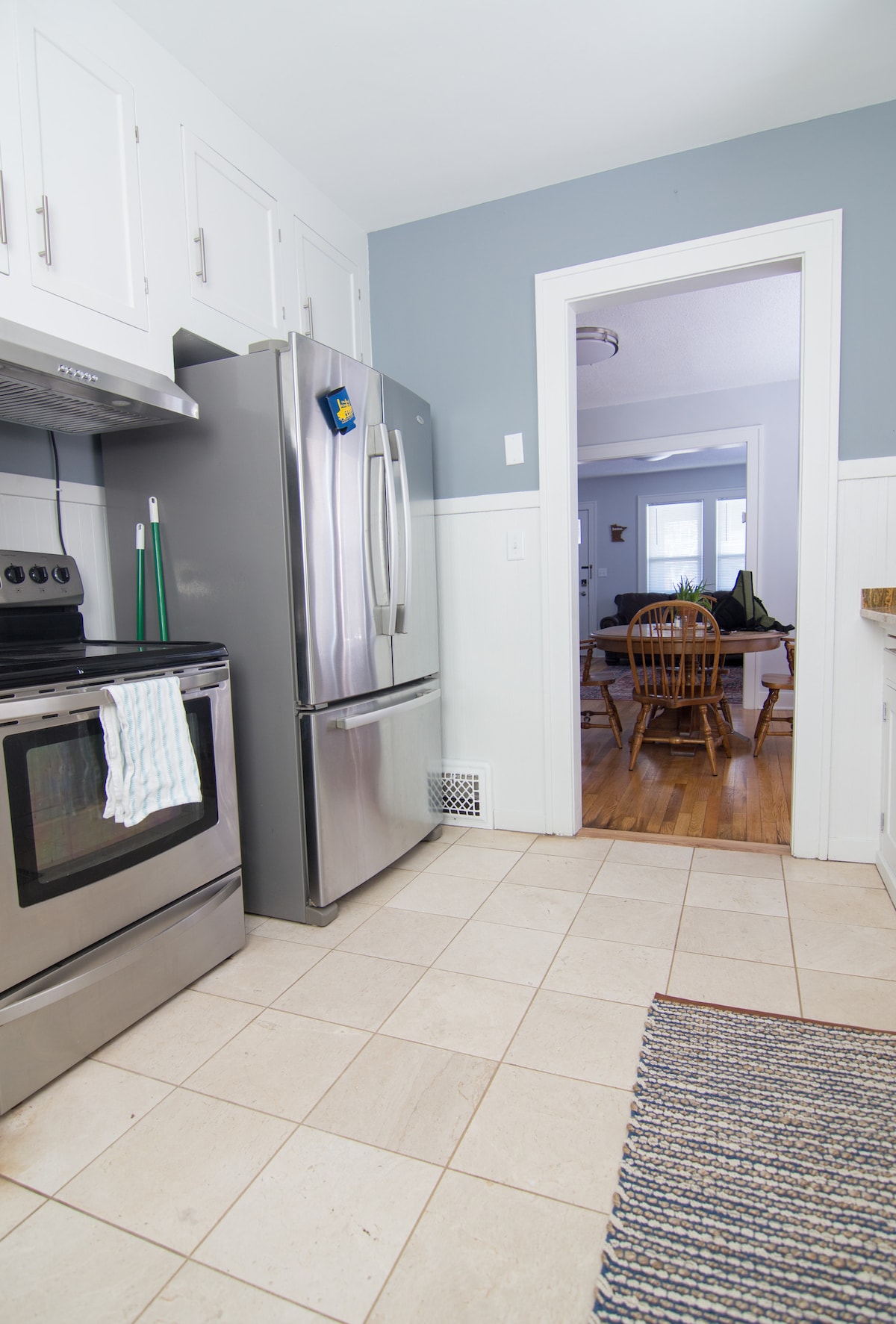 A modern kitchen features stainless steel appliances, including a refrigerator and an oven. Light-colored cabinets are positioned above the counter space, which is complemented by a tiled floor. A doorway leads to the adjacent dining area, creating an open and connected space.
