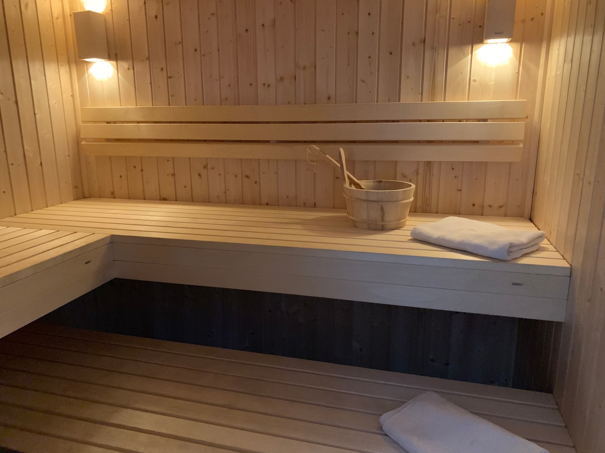 The sauna features wooden benches arranged in a minimalist design, with soft lighting coming from wall sconces. A wooden bucket and ladle are placed on one bench, while neatly folded towels are set on the other, creating an inviting and tranquil atmosphere.