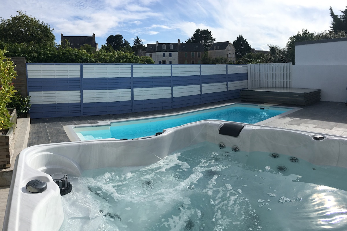 An outdoor area features a spacious hot tub adjacent to a swimming pool, both surrounded by modern fencing. Sunlight reflects off the water's surface, and greenery provides privacy. A wooden deck can be seen in the background, offering additional space for relaxation.