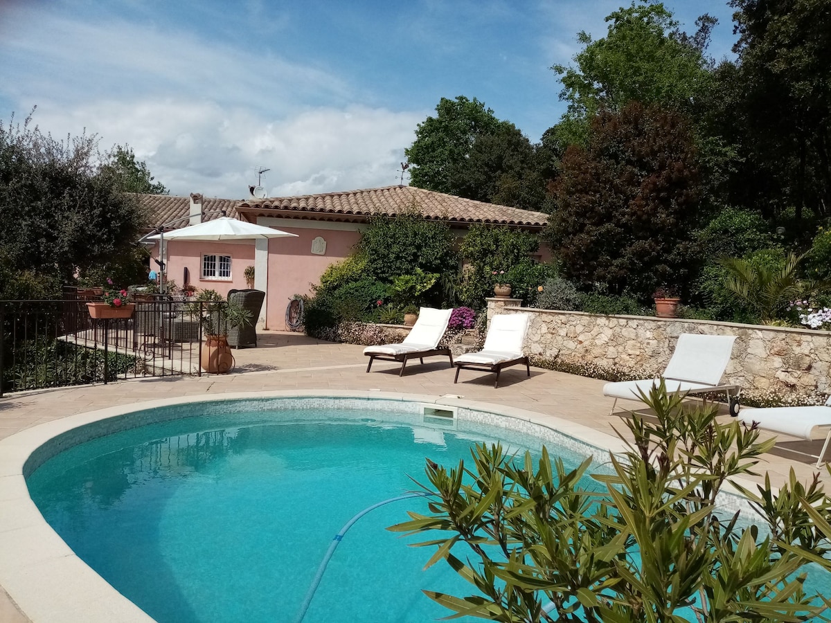 The image displays a tranquil outdoor pool surrounded by a stone terrace, featuring white lounge chairs positioned at the water's edge. Lush greenery is visible in the background, alongside a pink building with a tiled roof and well-maintained plants, creating an inviting outdoor space.