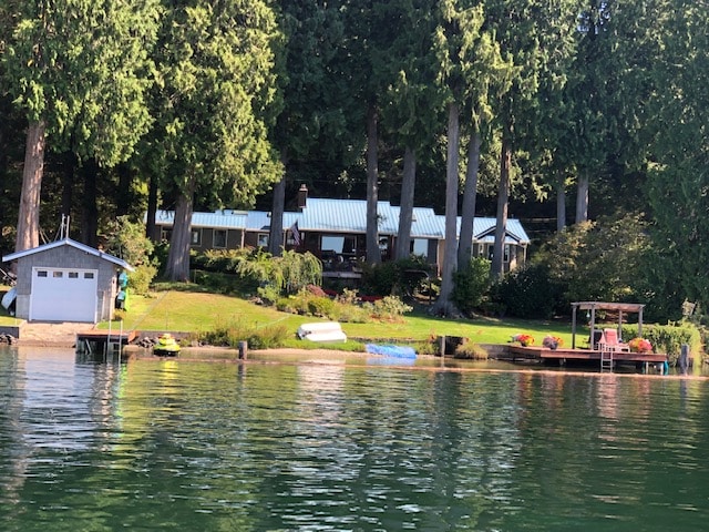 The cabin is nestled among tall cedar trees, featuring a blue metal roof and a spacious deck. A well-kept lawn with flowering plants enhances the outdoor space, while a couple of docks and colorful kayaks are visible along the water's edge.