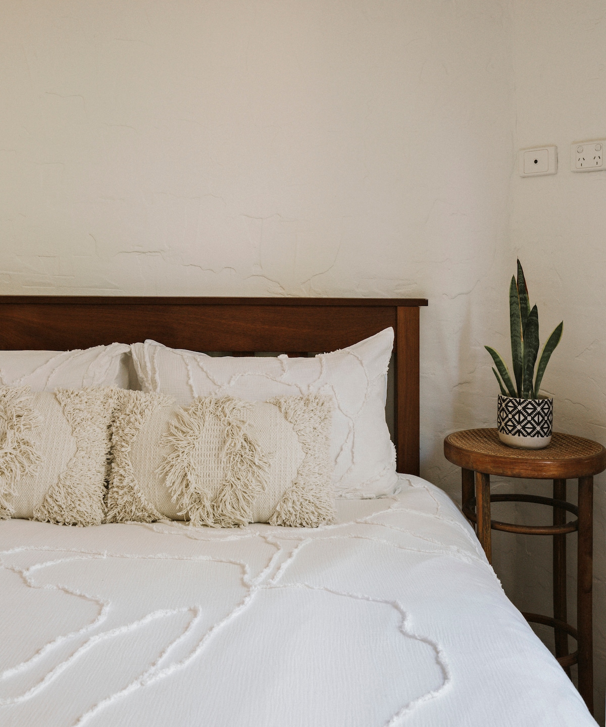A cozy sleeping area is presented with a spacious bed topped with textured white bedding. Two decorative pillows add a touch of comfort. Beside the bed, a small round table holds a potted plant, contributing a natural element to the room's serene ambiance.