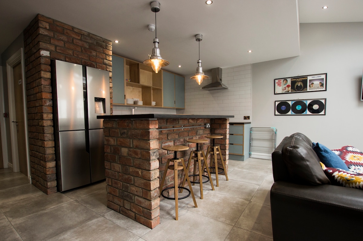 A modern kitchenette features a stainless steel refrigerator and a minimalist brick island with three wooden stools. The room is filled with natural light, and the design combines clean lines with a touch of warmth, creating a welcoming social space.