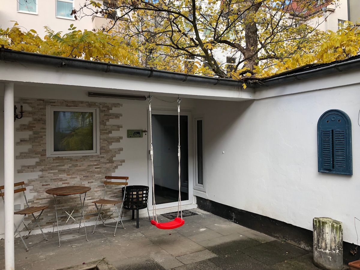 A covered terrace features a red swing hanging from the roof, alongside a small round table and two chairs. The wall is painted white with a stone accent, and a doorway leads to the guesthouse's interior.