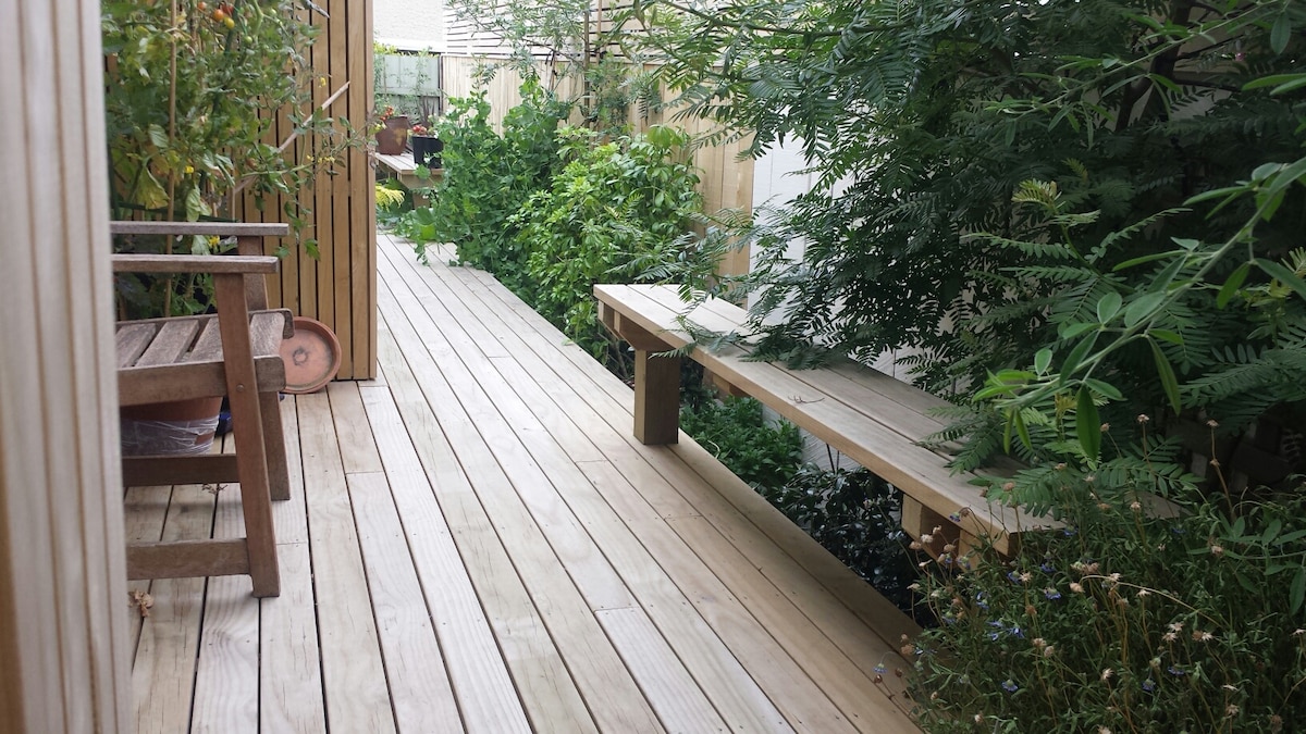A wooden pathway is flanked by lush greenery, creating a serene outdoor atmosphere. A bench is positioned along the path, providing a space to relax. The surrounding plants contribute to a sense of tranquility and connection with nature.