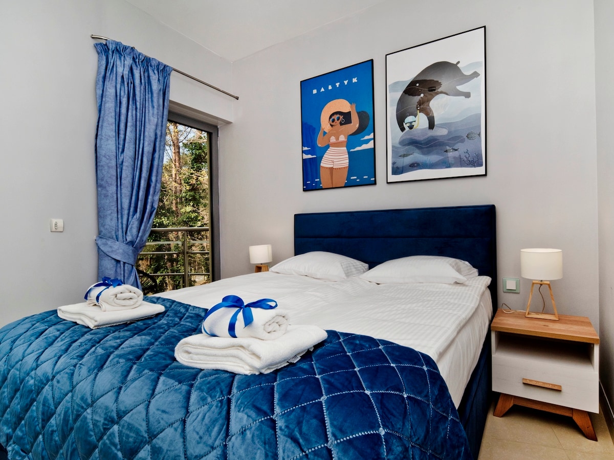 A stylish bedroom features a spacious bed with white linens and blue accents. Two vibrant art pieces adorn the walls above the bed. Soft light is provided by bedside lamps on each side, and a window with blue curtains offers a view of the outdoors.