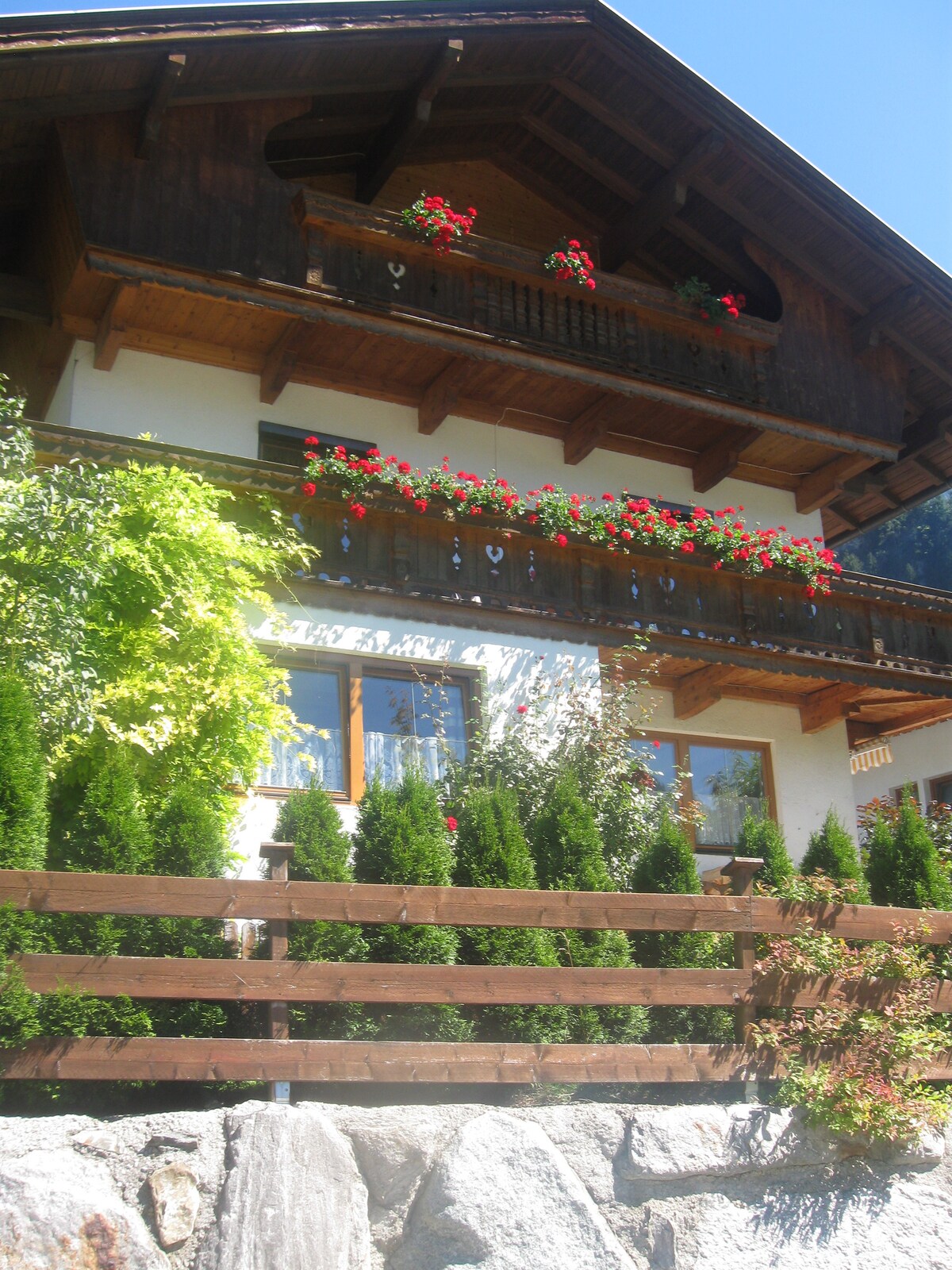 A traditional Zillertal house features wooden balconies adorned with vibrant red flowers. Lush greenery lines the front, complemented by a rustic wooden fence and a stone foundation, providing a welcoming entrance against a clear blue sky.