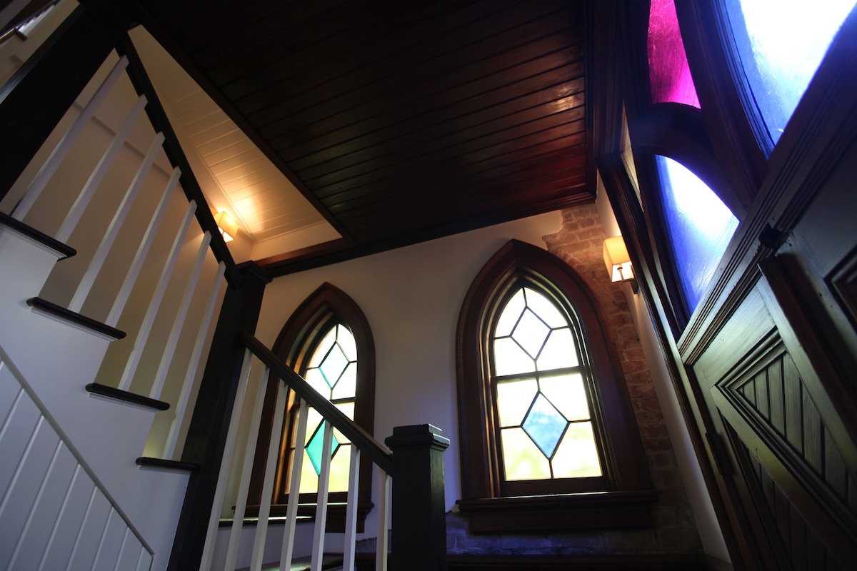 The interior staircase is framed by original arch-top windows with stained glass in rich hues, allowing soft light to filter through. Wooden paneling and a high ceiling enhance the sense of space. The stairs have a simple railing, leading to the upper level.