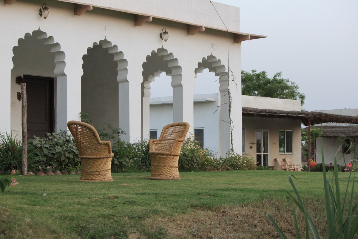 A quaint outdoor seating area features two woven chairs positioned on a well-maintained lawn. Arched architectural elements frame the entrance to the farmhouse, with lush greenery and various plants in the background. The scene conveys a peaceful and inviting outdoor space.