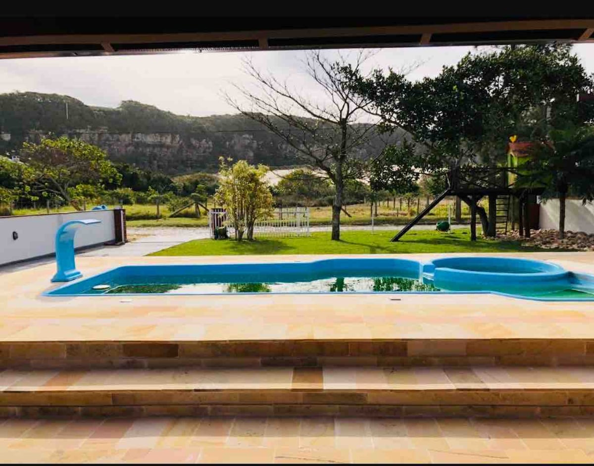 A serene pool area is viewed from an outdoor space, featuring a crisp blue pool surrounded by a stone deck and lush green grass. Natural elements like trees and hills in the background create a tranquil setting, ideal for relaxation.