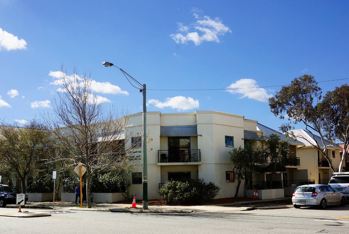 An exterior view of a light-colored two-story building is shown, featuring balconies and large windows. Surrounding greenery includes small trees and shrubs, contributing to a pleasant streetscape. A clear sky with few clouds is visible above the structure.