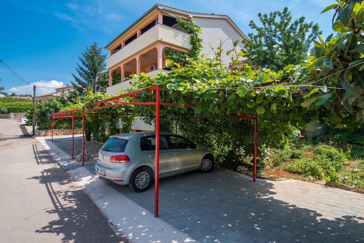 A shaded parking area is located under a trellis adorned with green vines, accommodating a single parked car. The surrounding landscape includes well-maintained greenery and a glimpse of the apartment above, situated in a peaceful neighborhood.