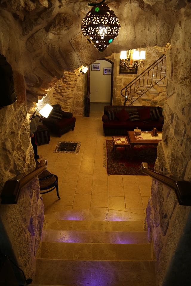 The image showcases a stone staircase leading down to a warmly lit common area. Traditional seating arrangements are visible, along with a decorative table in a richly adorned space. An ornate lantern hangs from the ceiling, casting a soft glow across the tiled flooring.